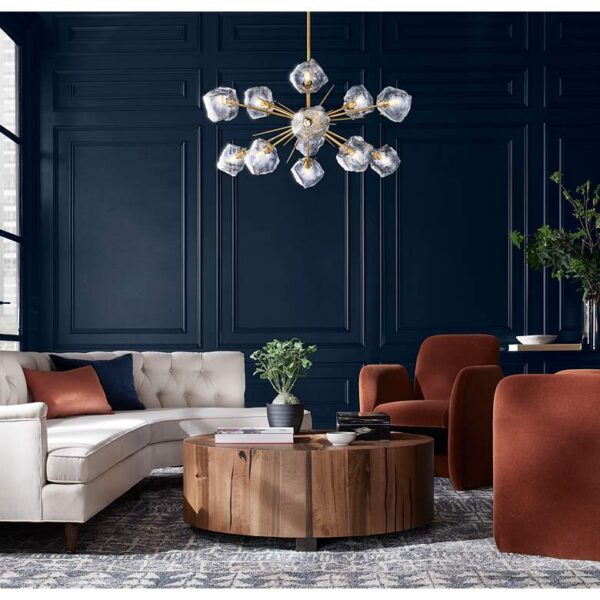 A golden bronze chandelier with clear ice glass globes hanging from the ceiling. The chandelier is suspended from a chain and has a decorative finial at the top. The chandelier is located in a living room with a couch, coffee table, and chairs.