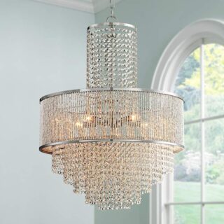 A product image of a Pioggia Chrome 23 1/2 Wide Crystal Chandelier. The chandelier is made of crystals and has a round base. It is hanging from the ceiling in a living room.